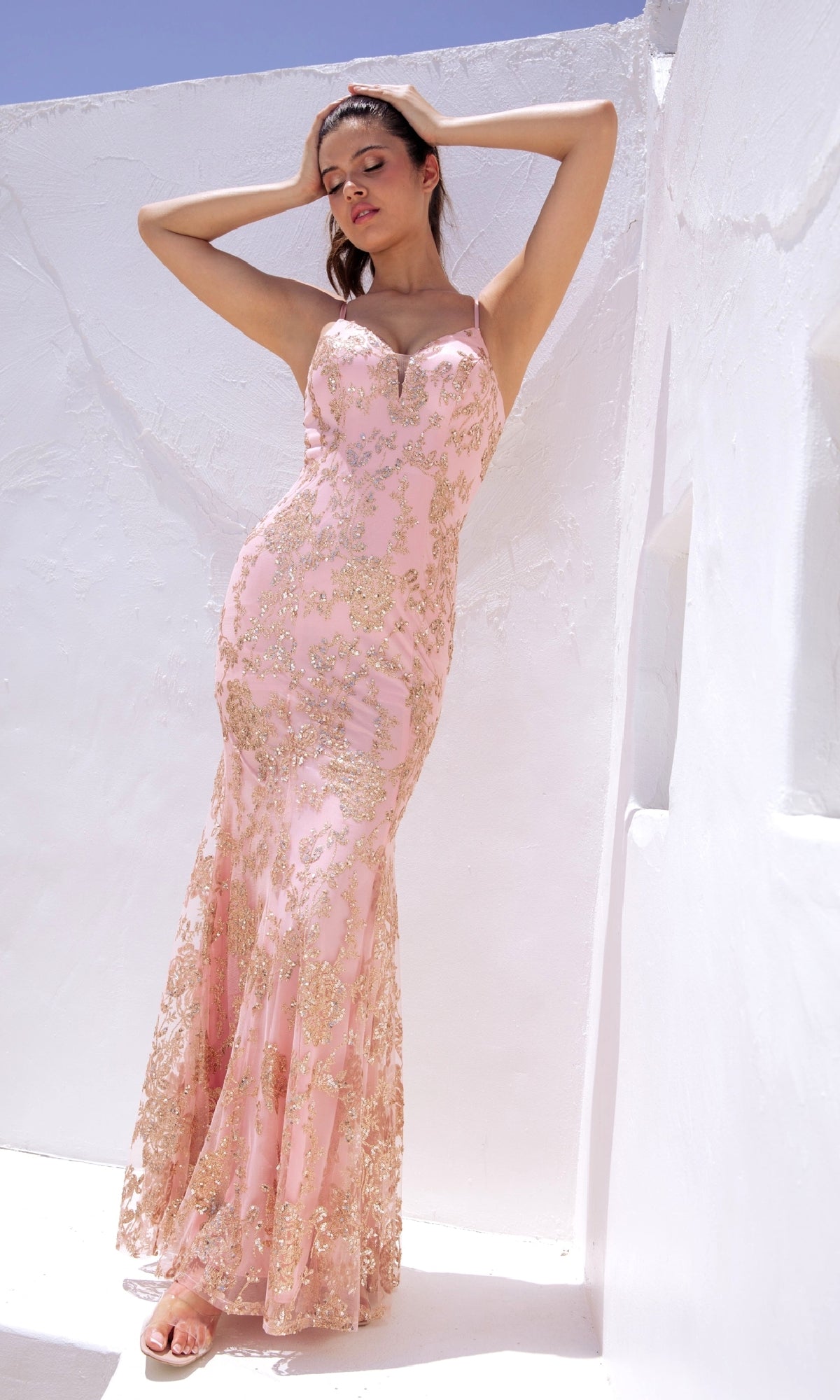 A woman stands against a whitewashed wall in bright sunlight, wearing the Eureka 9788 Strapless Long Glitter-Print Prom Dress—a fitted blush pink mermaid gown with gold sequin embellishments.